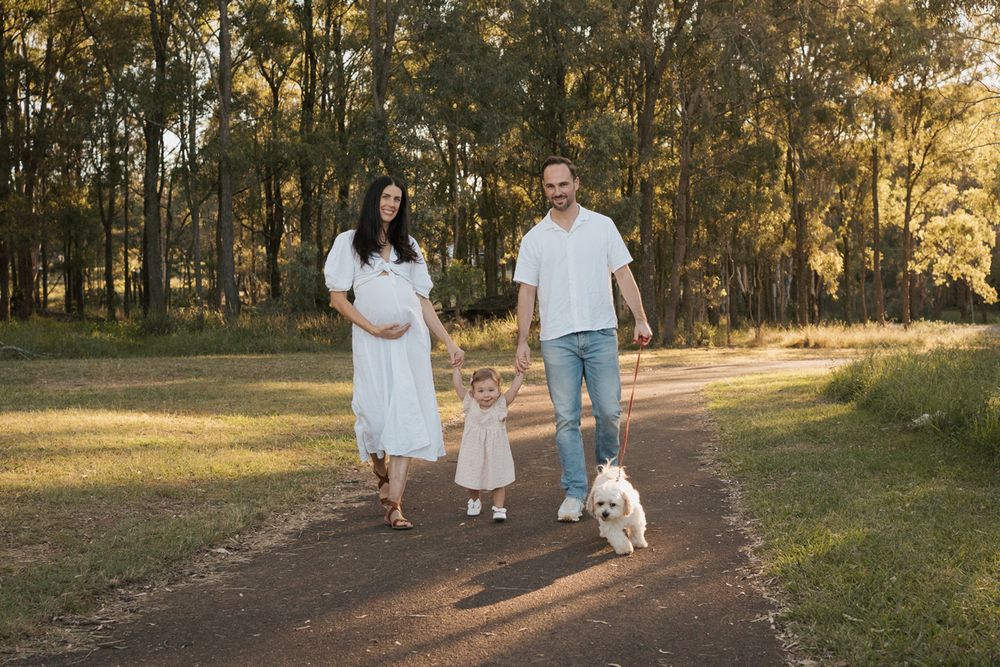 Pregnant woman in white dress walks with partner, toddler, and white dog on a sunny park path surrounded by trees in Rouse hill photoshoot