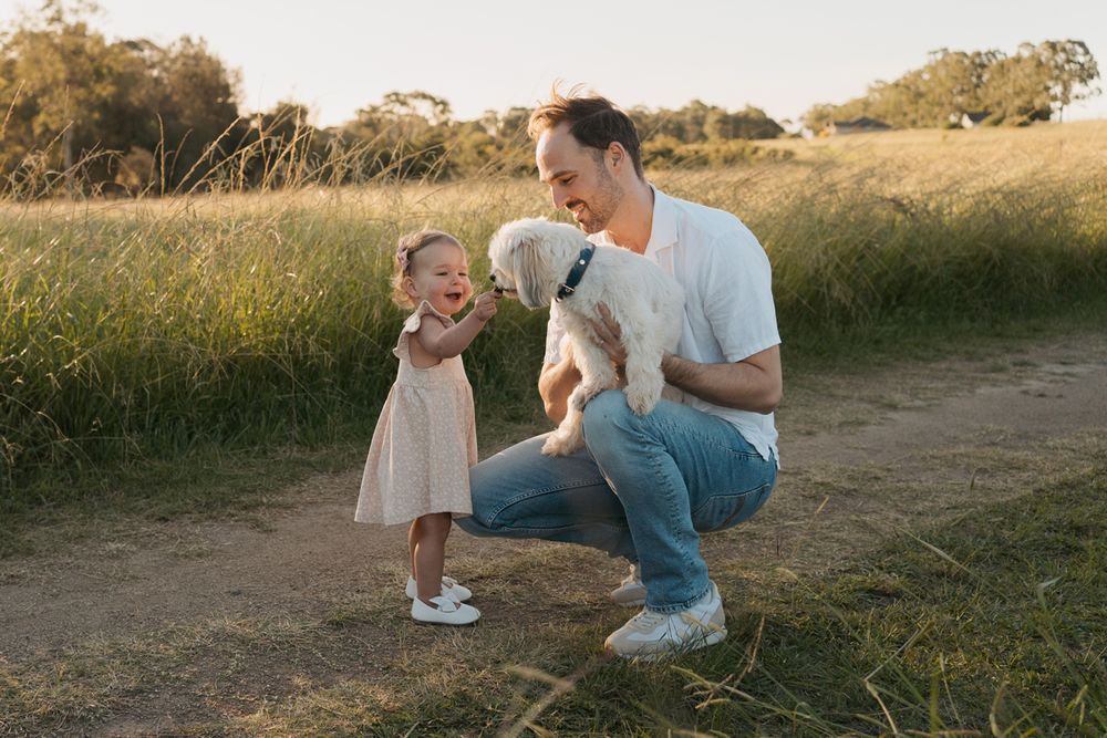 Father crouching with toddler girl and white puppy on a sunny dirt path surrounded by green grass in rouse hill family photoshoot with dog