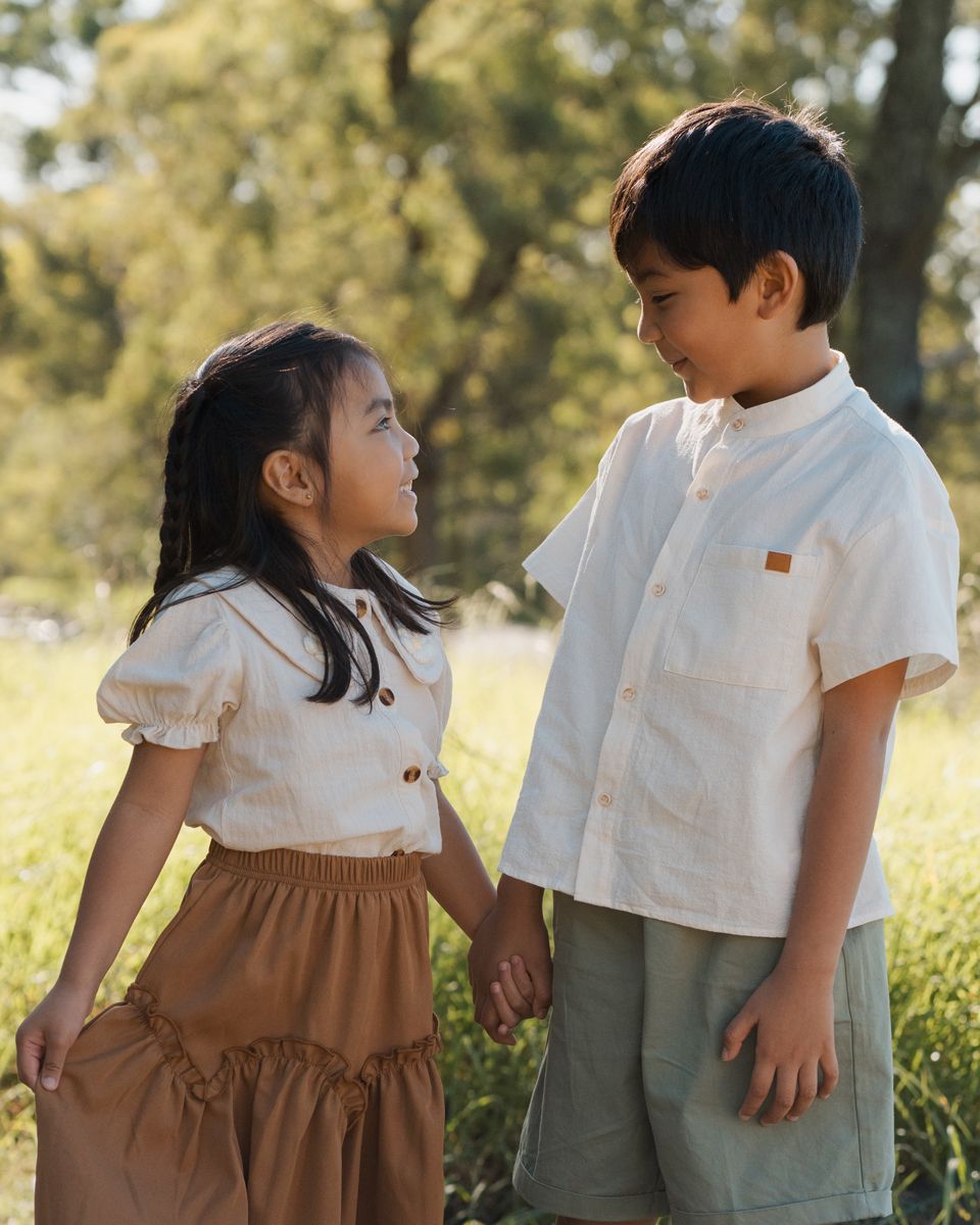 Two young siblings in matching linen outfits hold hands in a sunlit park, smiling at each other during a family photoshoot in Rouse hill