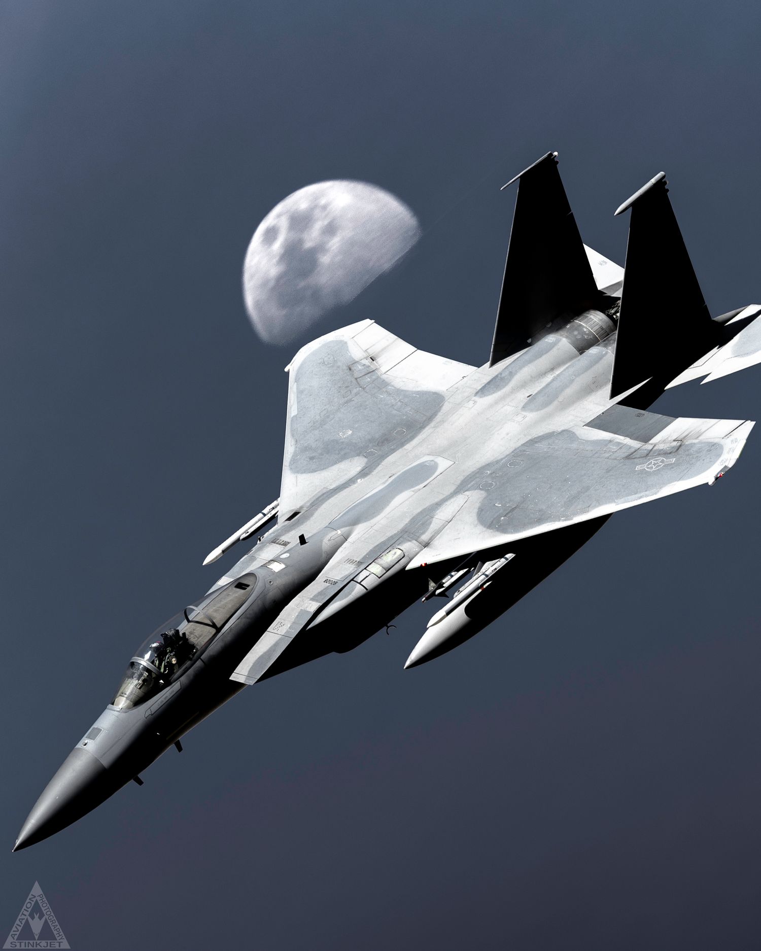 An F-15C Flying against a dark blue sky background with the moon hanging just off of the right wingtip in the background
