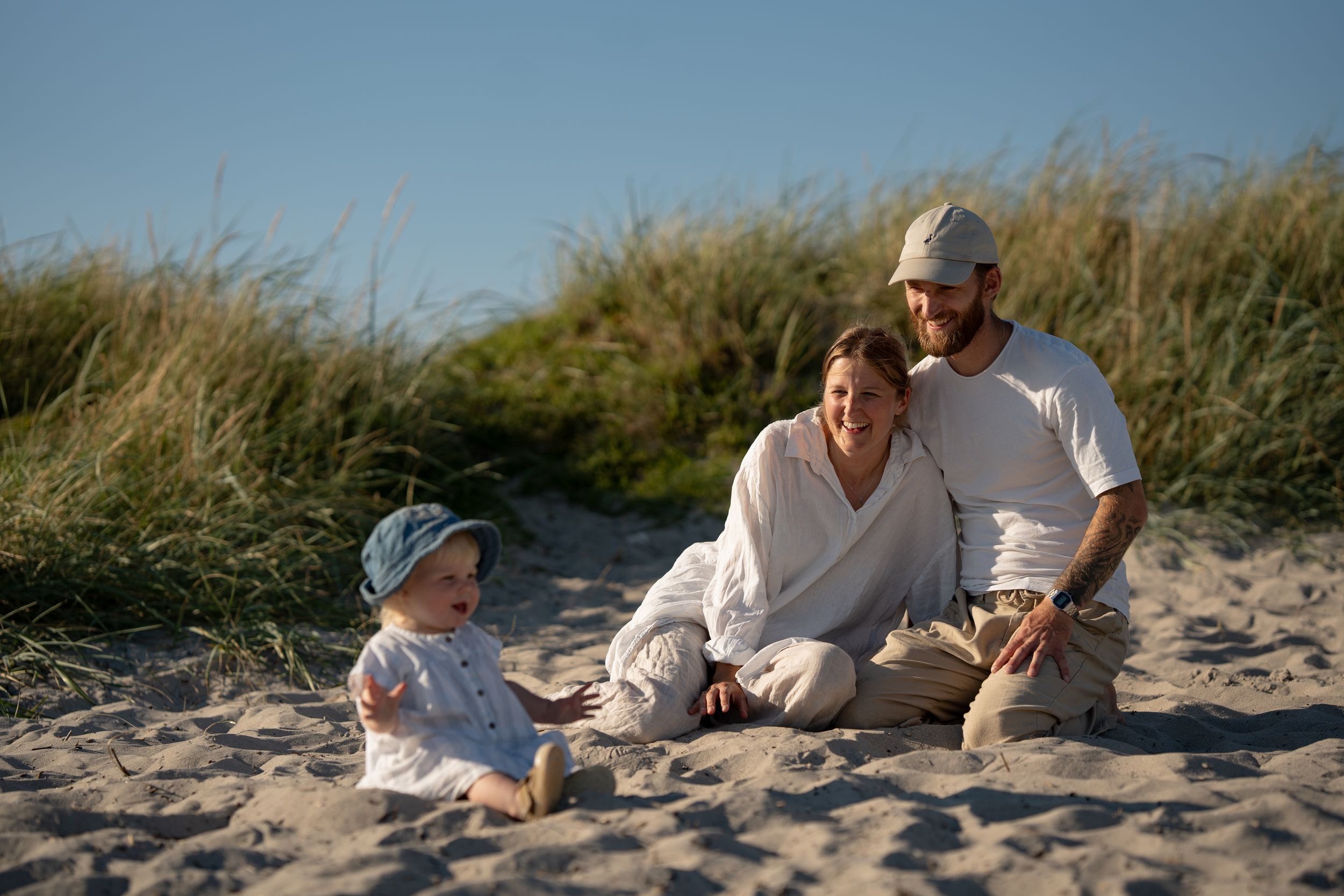 En mamma och pappa i ljusa kläder sitter på en strand och tittar på sin dotter som leker i sanden. De skrattar.