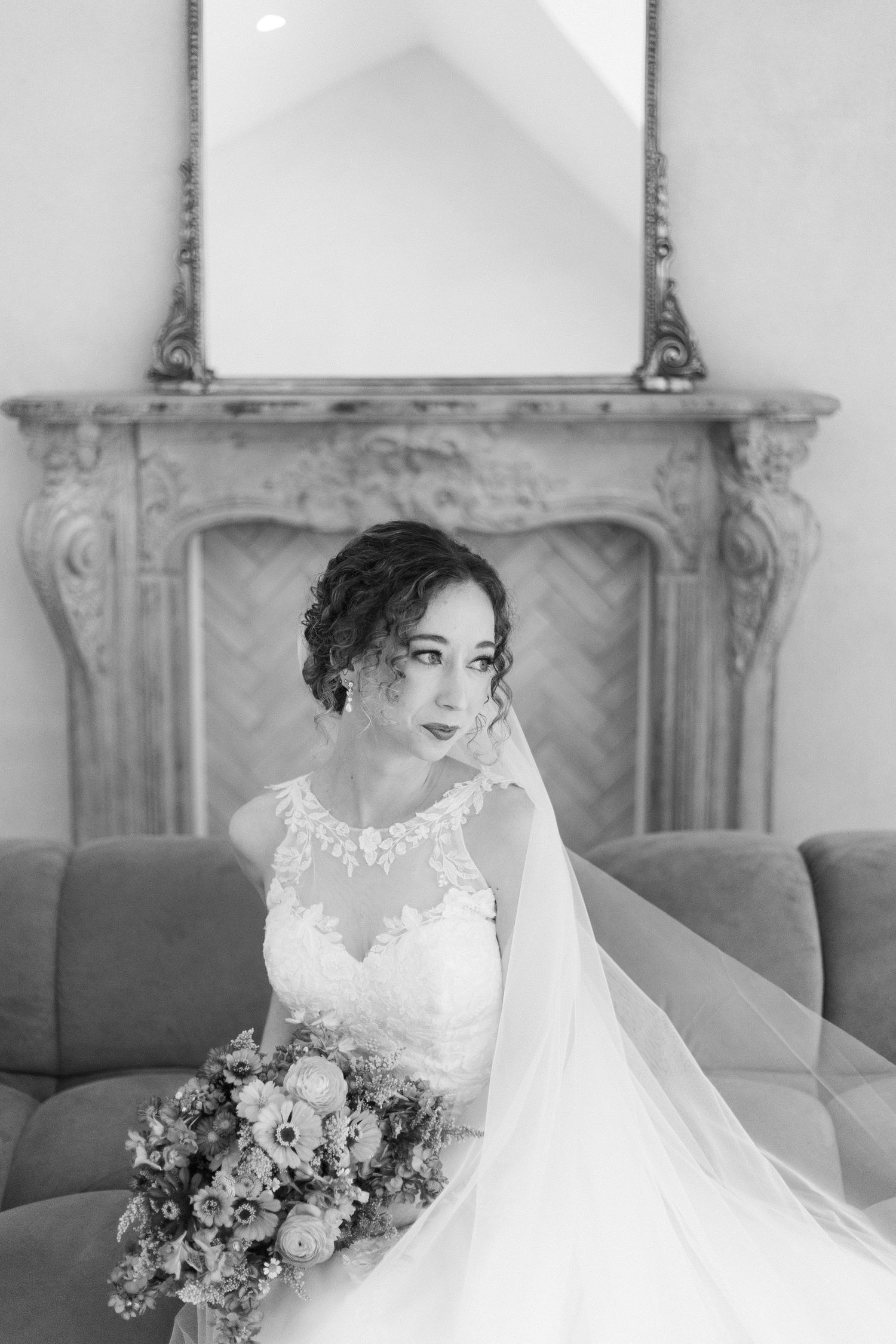 Black and white bridal portrait of a bride in lace gown holding bouquet, seated before ornate fireplace with veil flowing.