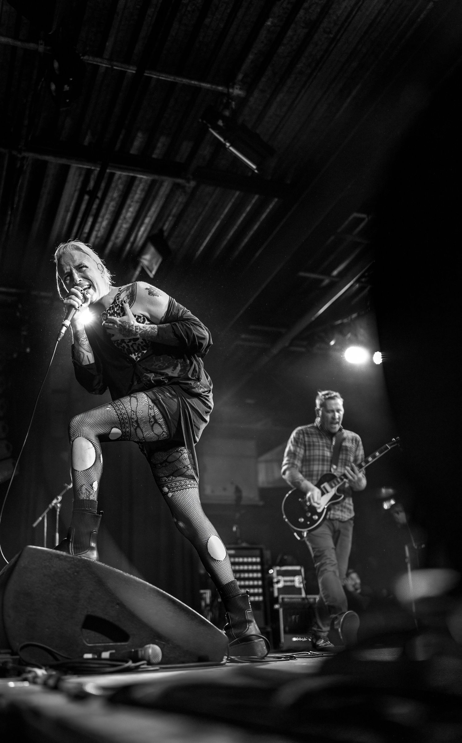 Black and white photo of a female punk rock singer performing on stage with a guitarist playing Les Paul in the background.