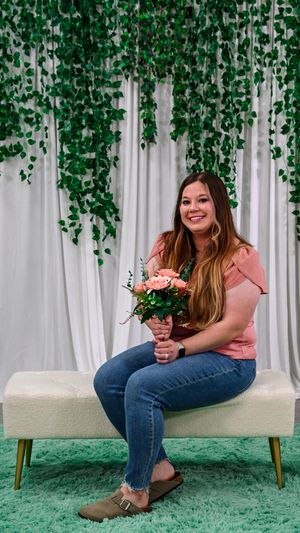 Woman in pink top and jeans sits on white bench holding pink roses with green ivy backdrop.