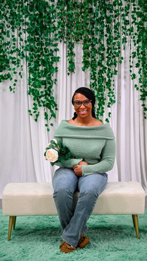 Smiling woman in green off-shoulder sweater and jeans holds white rose on bench with ivy backdrop.