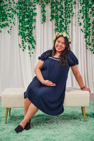 Pregnant woman in navy dress with floral crown poses on bench with green ivy backdrop.