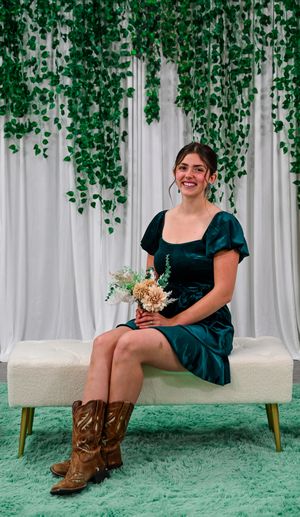 Woman in teal dress and cowboy boots sits on white bench holding bouquet with green vine backdrop.