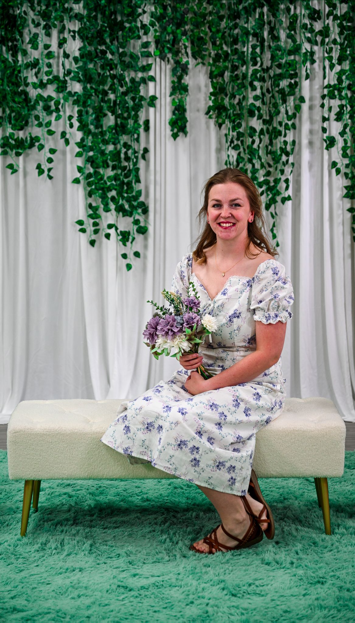 Woman in floral dress holding purple bouquet sits on white bench with green ivy backdrop.