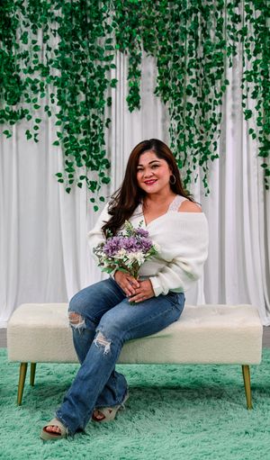 Woman in white sweater and jeans holds flower bouquet on bench with green ivy backdrop.