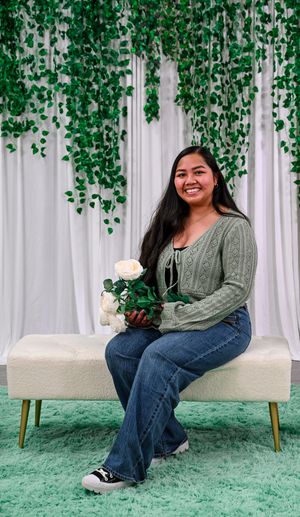 Smiling woman in sage cardigan and jeans holds white roses on bench with ivy backdrop.
