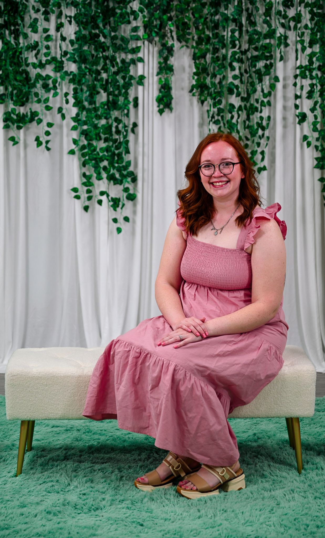 Smiling woman in pink maxi dress sits on white bench with hanging greenery backdrop.
