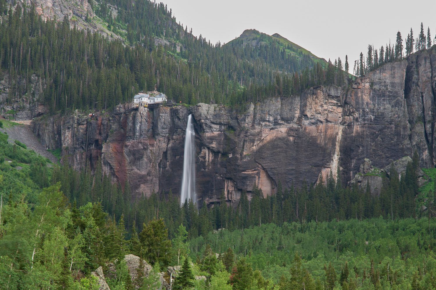 A dramatic tall waterfall plunges down rugged rocky cliffs surrounded by dense pine forest and mountains. Telluride, CO Bridal Veil Waterfal