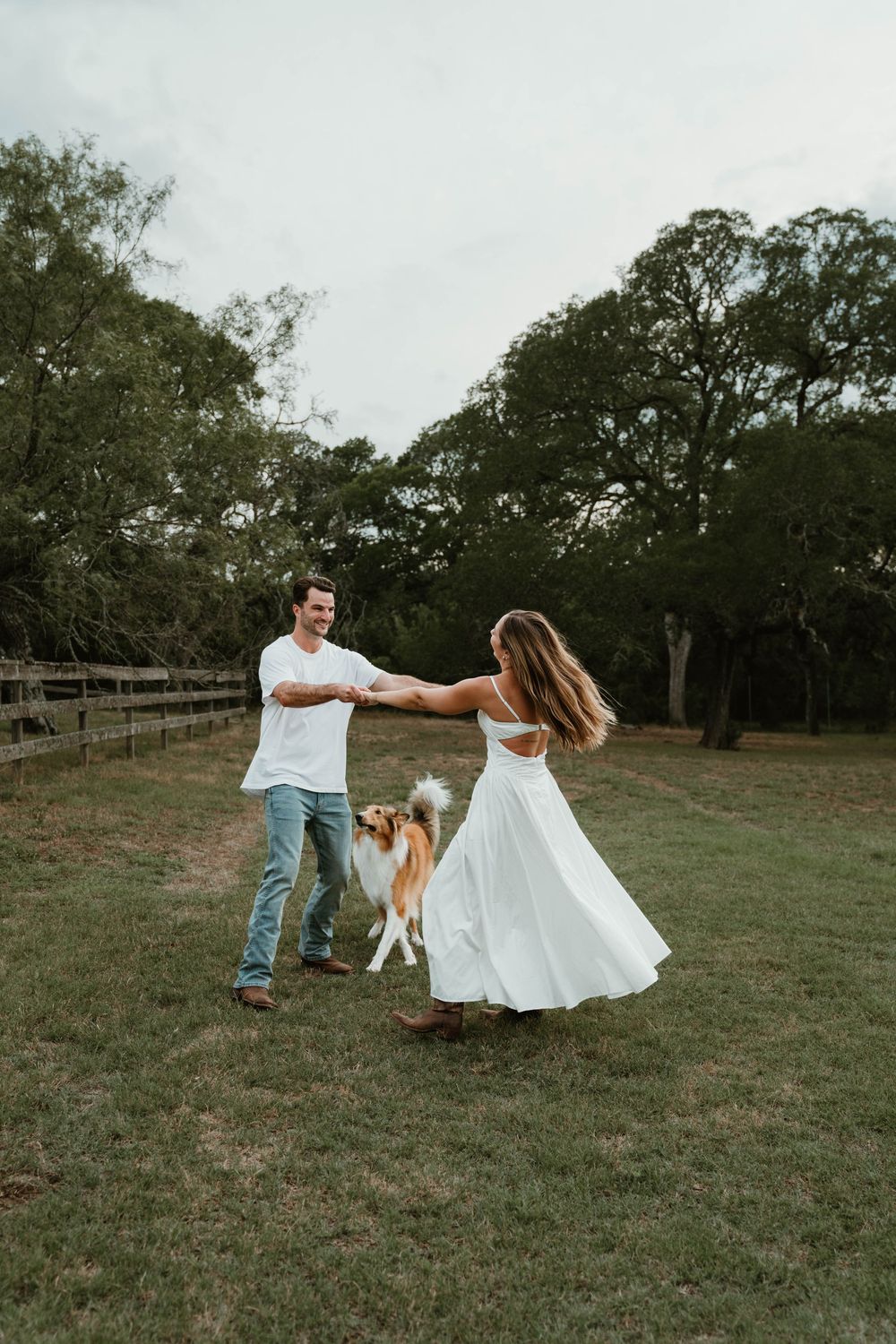 Couple dancing in a field with a Collie dog, woman in white dress, man in jeans, surrounded by trees.