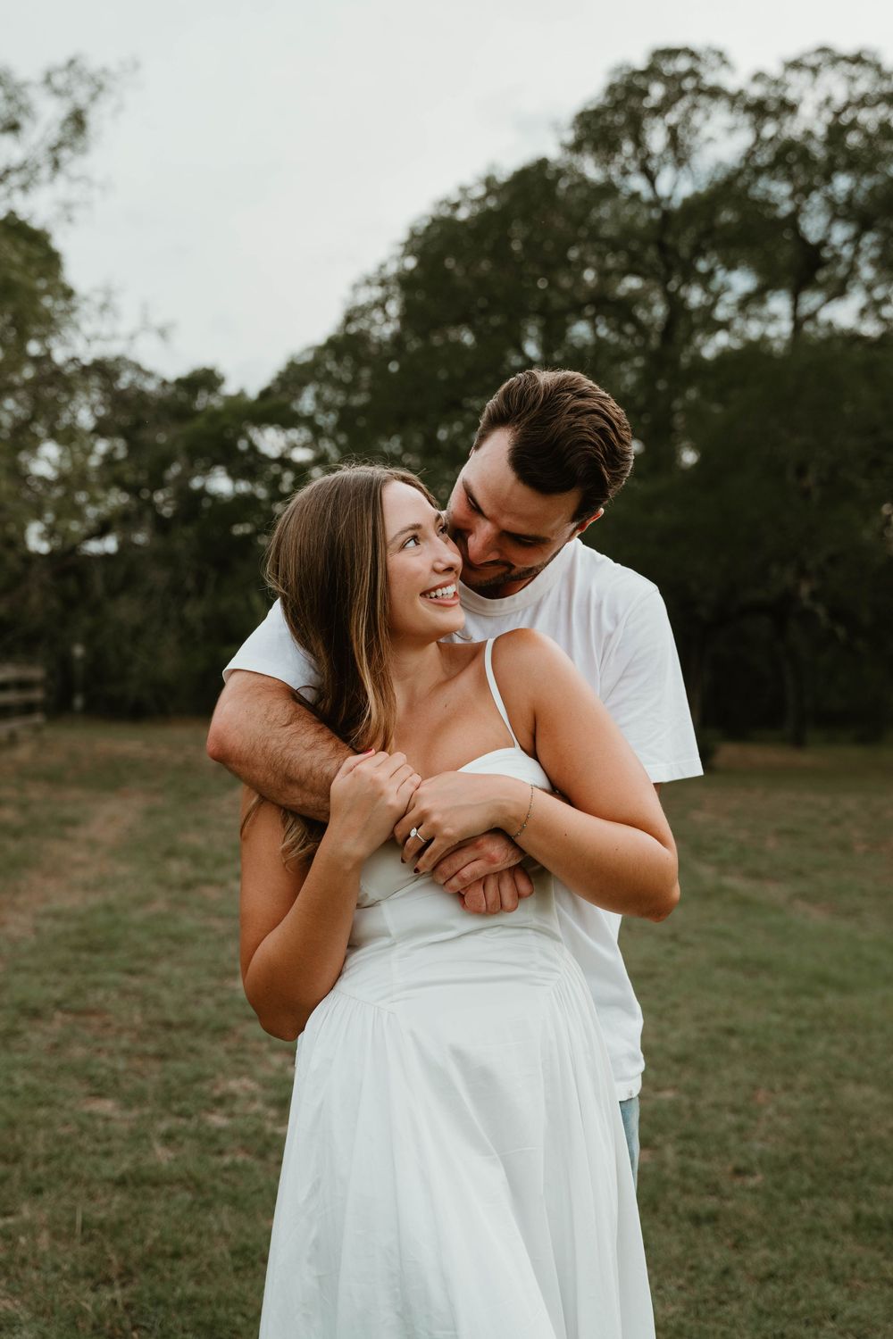 A couple embraces in a green field, woman in white dress smiling as man hugs her from behind near trees.