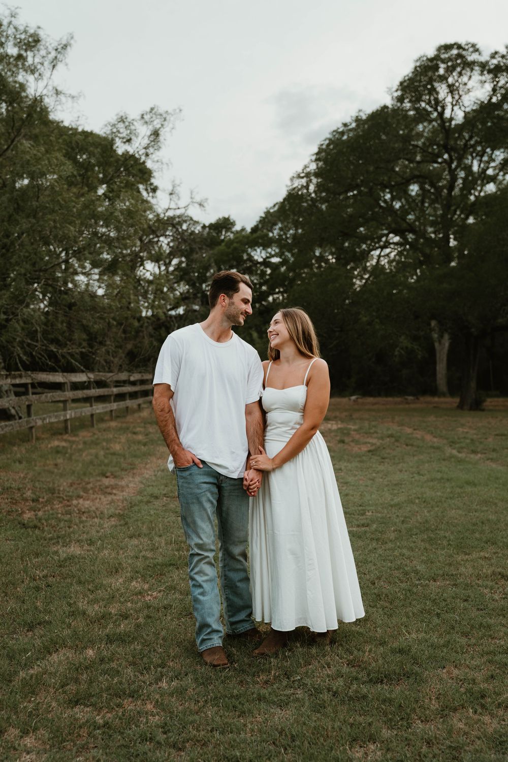 A couple holding hands and smiling at each other in a green field, wearing white outfits with trees in the background.