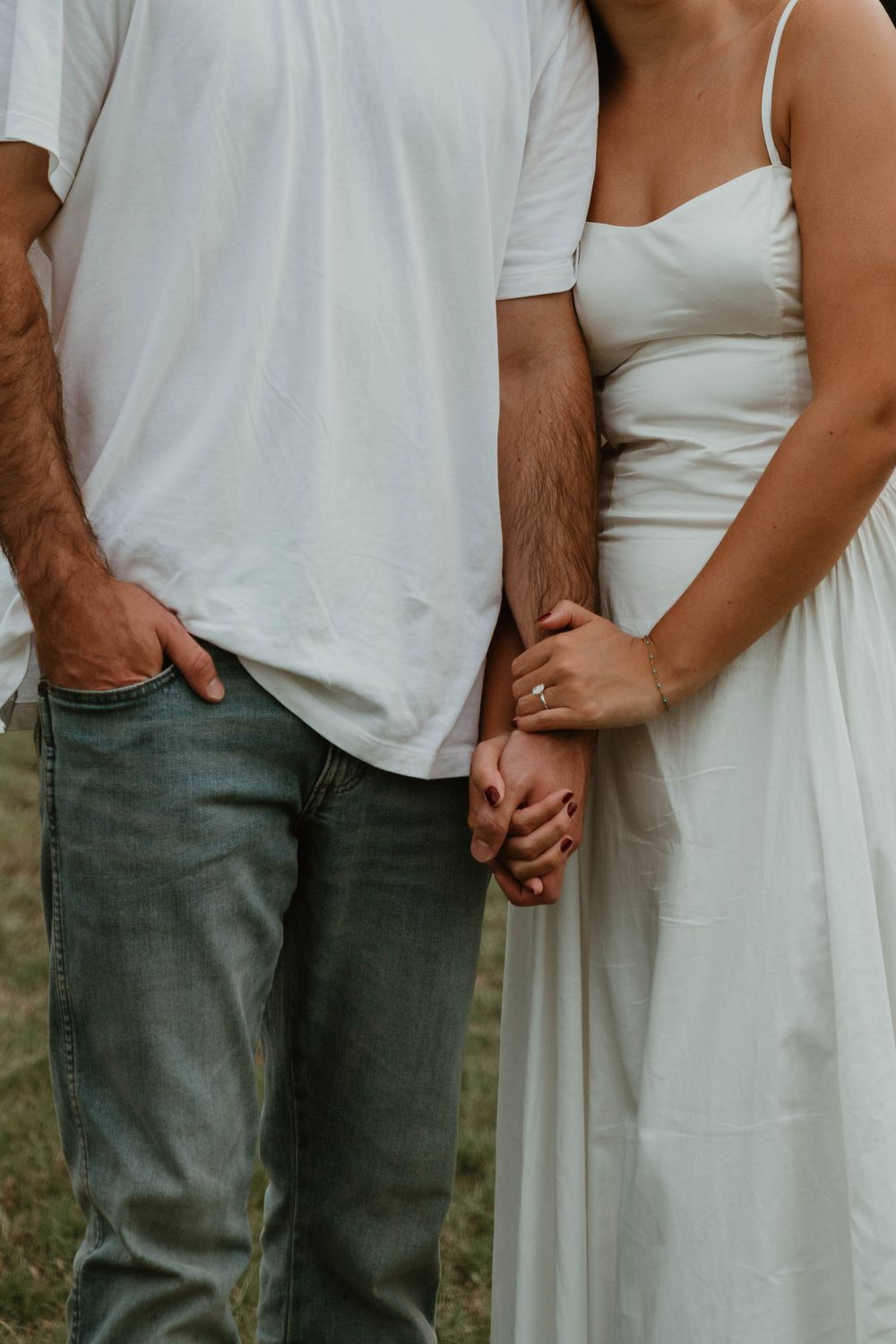 Couple holding hands, woman in white dress with engagement ring, man in white tee and jeans, romantic outdoor photo.