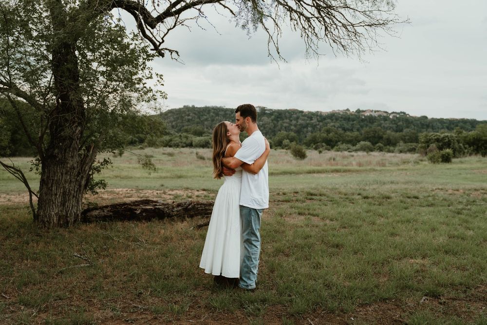 A couple embraces romantically in a lush green field beside a large tree with rolling hills in the background.