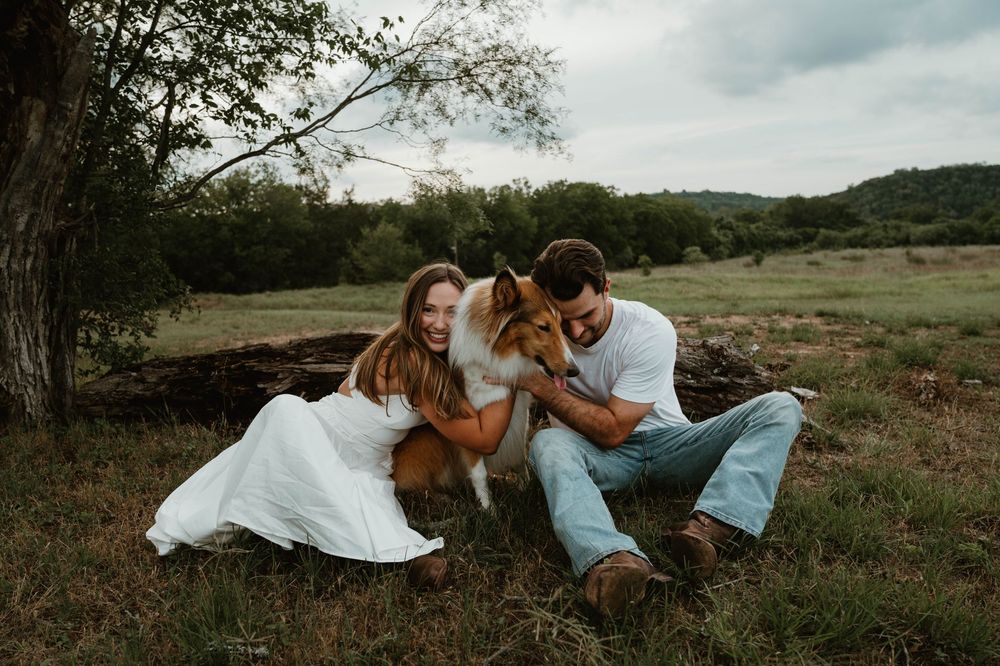 Couple in white outfits sitting on grass with a Collie dog in a scenic green field.