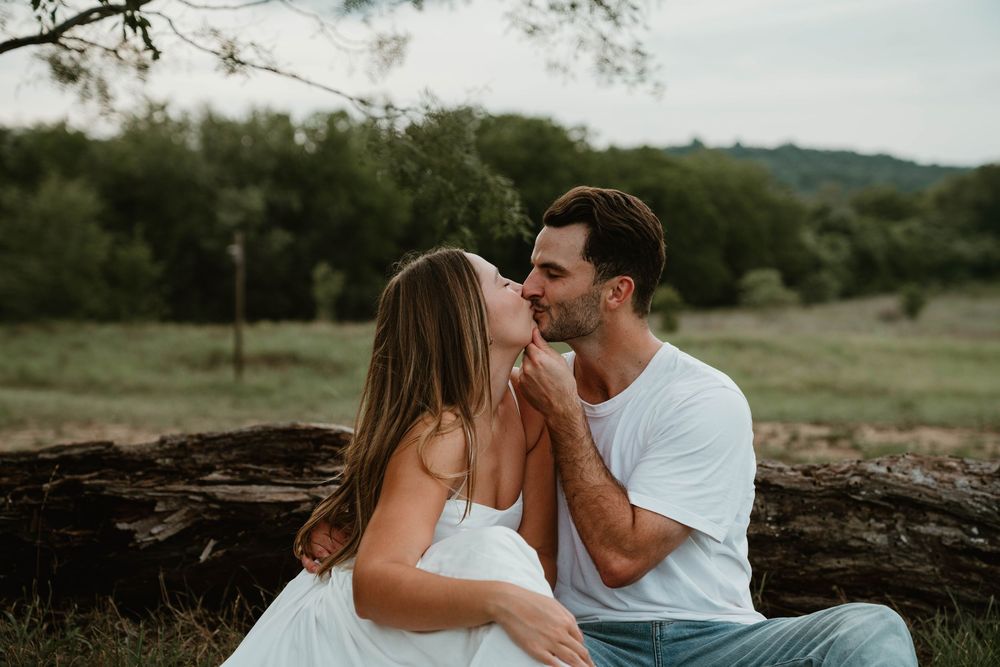 Couple sharing a romantic kiss outdoors, sitting on a log surrounded by lush green trees and fields.