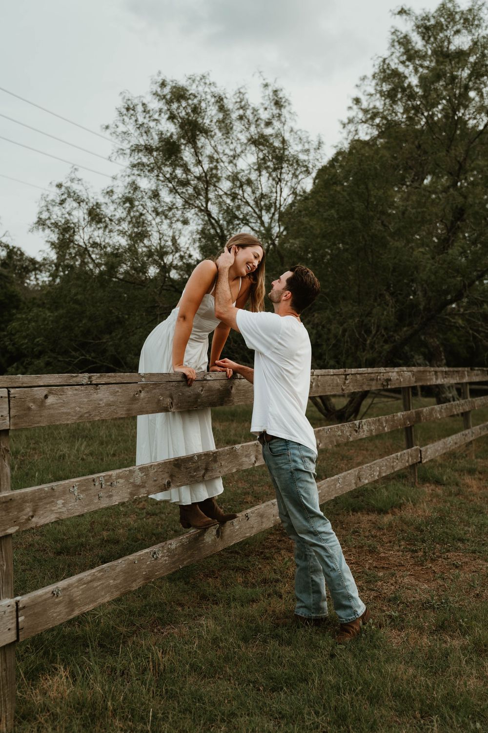 A couple shares a romantic moment by a wooden fence, woman sitting on top while man gazes up at her.