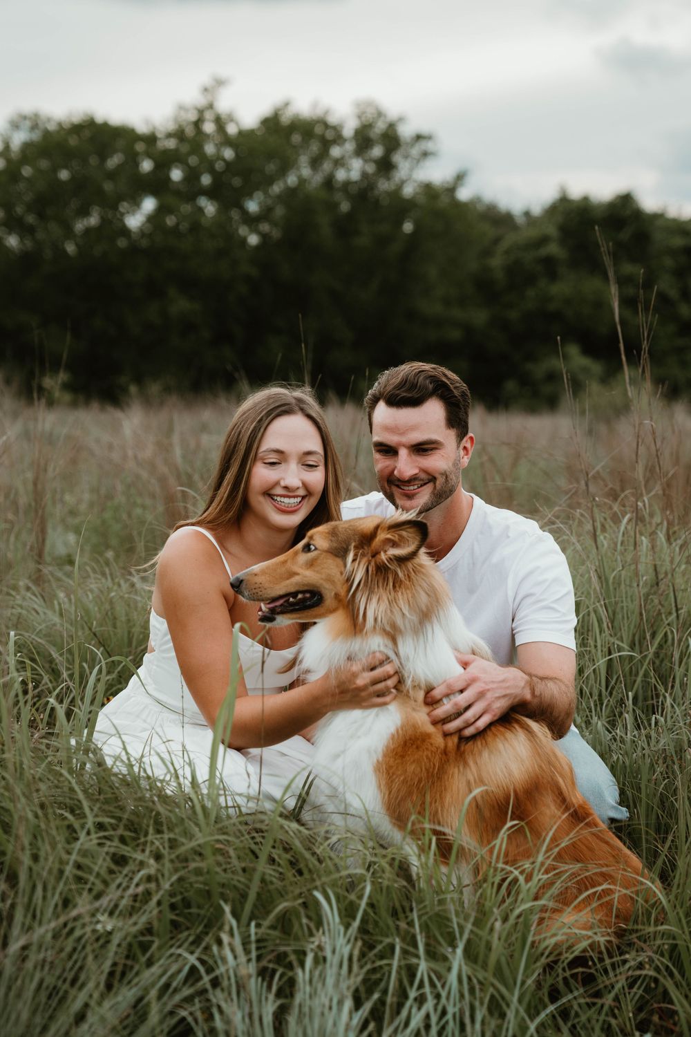 A smiling couple in white sits in tall grass with their golden Collie dog in a lush outdoor field.