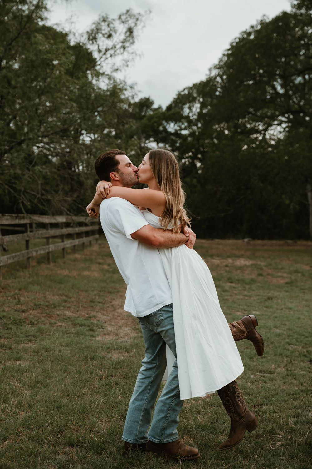 Couple sharing a romantic kiss in a green field, man lifting woman in white dress and brown boots.