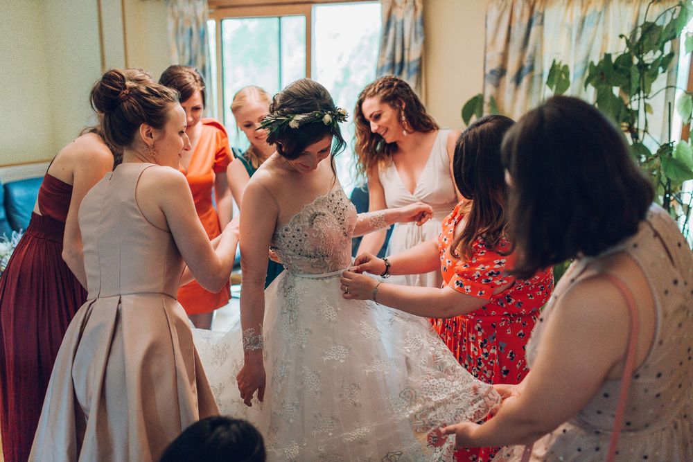 Bridesmaids helping bride button up her lace wedding dress during bridal preparations.