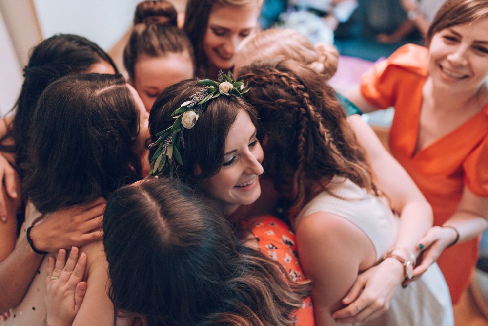 Bride wearing floral crown shares joyful group hug with bridesmaids before wedding.
