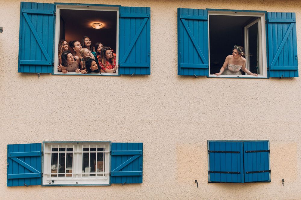 Wedding party peers out of open blue shuttered windows of a French building.