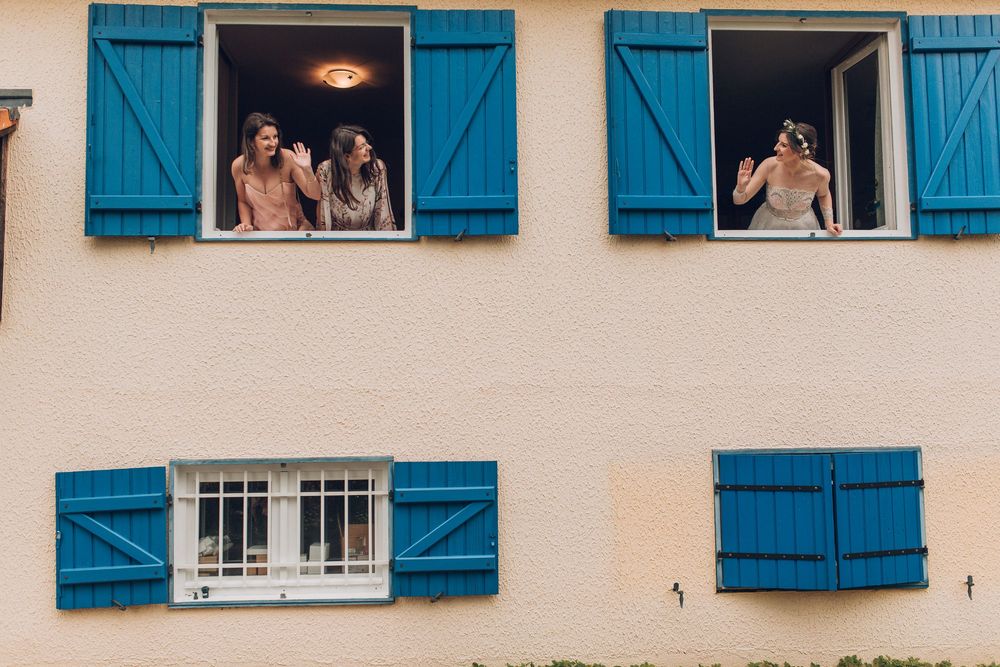 Bride and bridesmaids lean out of blue shuttered windows waving before the ceremony.