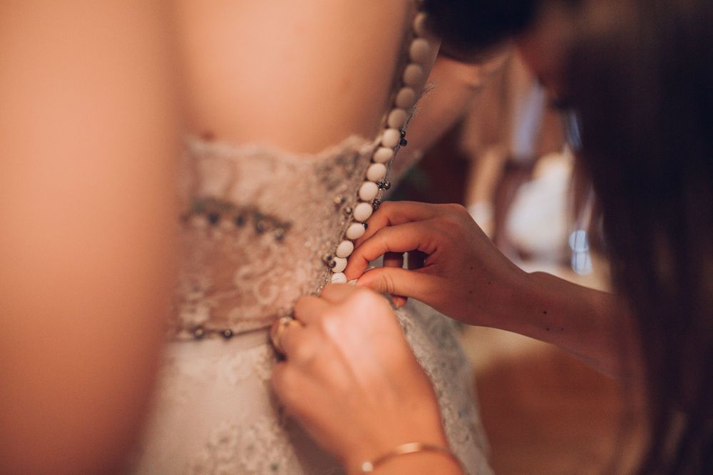 Close-up of bridesmaid carefully fastening pearl buttons on back of lace wedding gown.