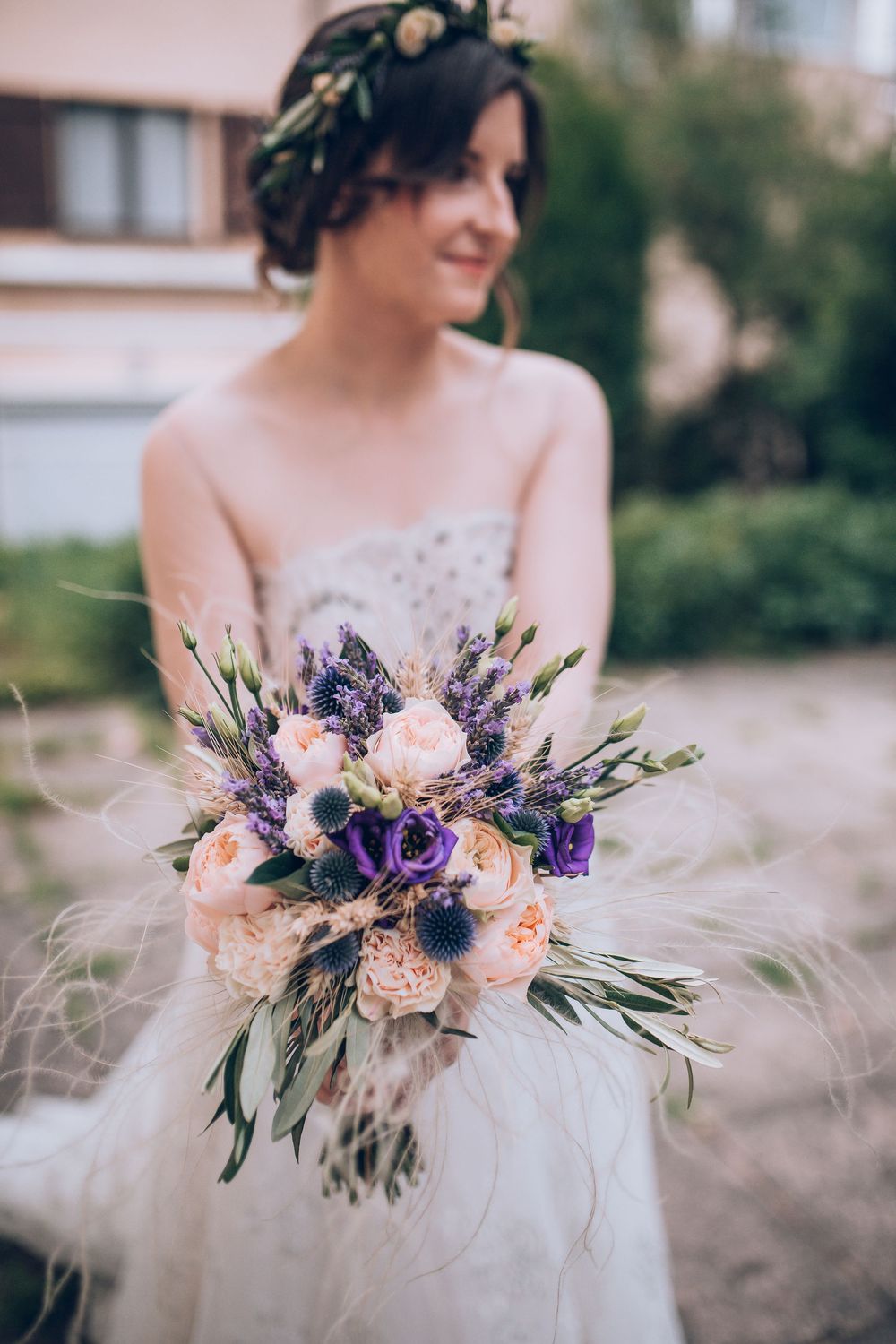 Bride in floral crown holds a stunning purple and blush wedding bouquet outdoors.