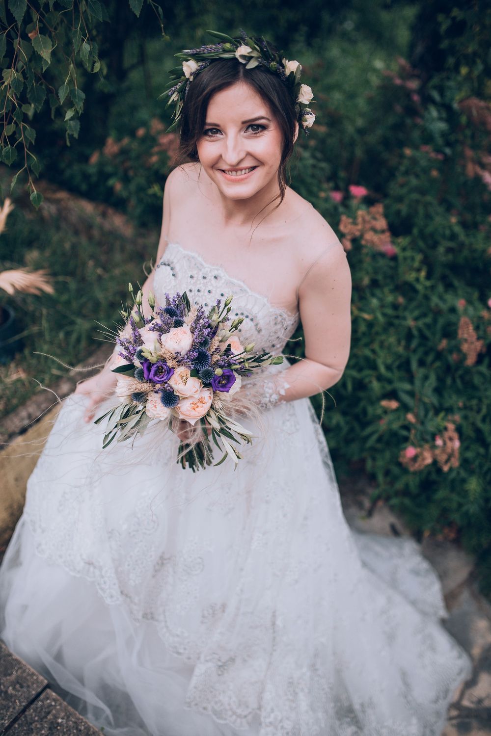Smiling bride in floral crown holds purple and blush bouquet in lush garden.