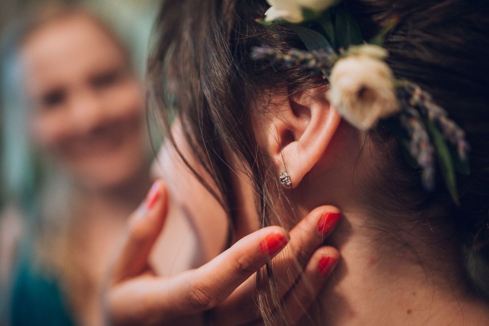 Close-up of bride's ear with diamond stud earring, adorned with flowers in hair, as bridesmaid helps prepare.
