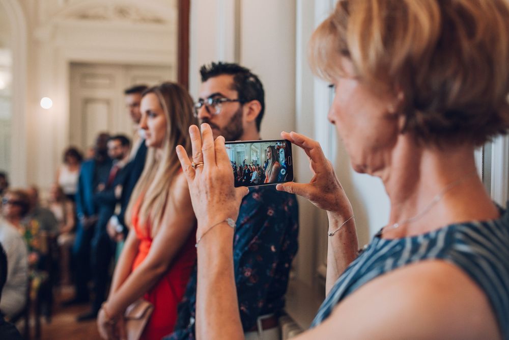 Wedding guest photographs ceremony on smartphone while attendees gather in elegant hall.