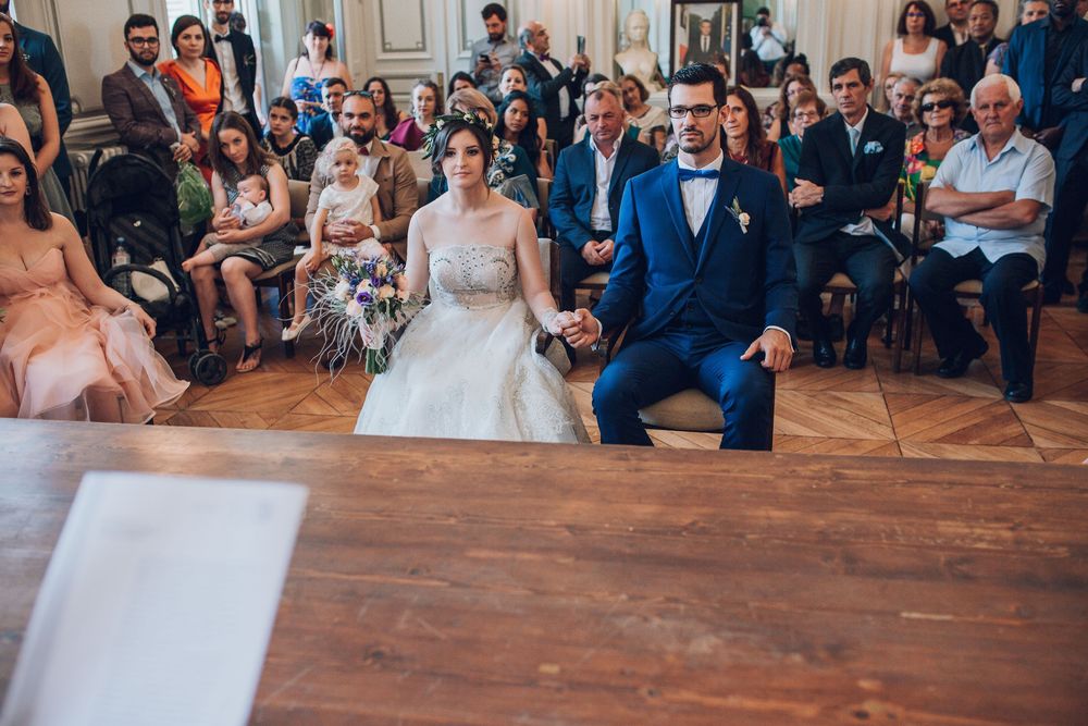 Bride and groom hold hands before officiant during civil wedding ceremony with guests seated behind.
