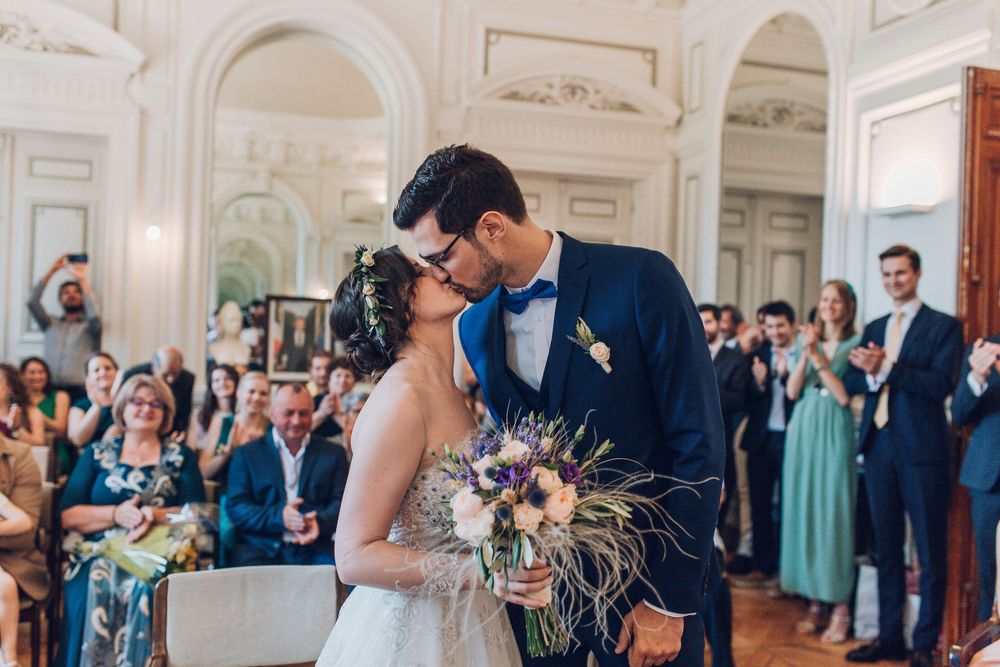 Newlyweds share first kiss as married couple while guests applaud in elegant French wedding hall.