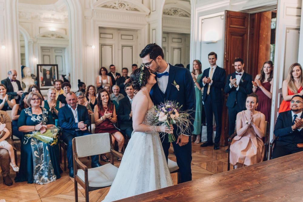 Bride and groom kiss during wedding ceremony as guests stand and applaud in ornate reception hall.