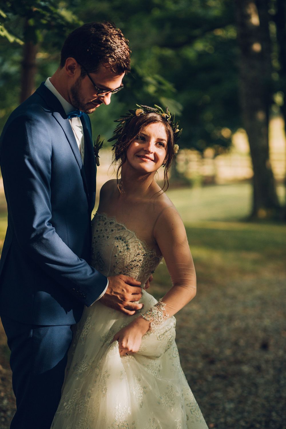 Smiling bride wearing floral crown embraces groom in blue suit in sunlit garden setting.