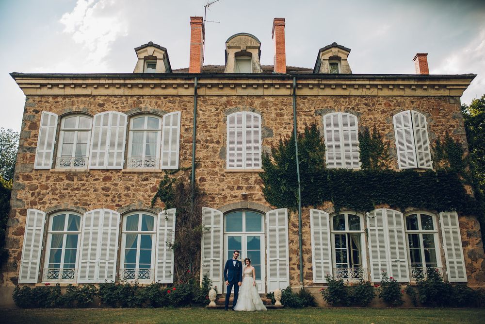 Wedding couple standing before a grand stone French chateau surrounded by lush ivy and gardens.