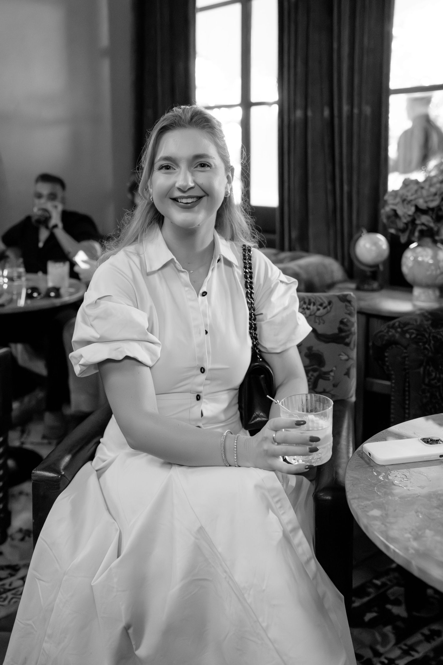 Smiling young woman in white dress holding a drink, seated in an elegant interior, black and white photo.