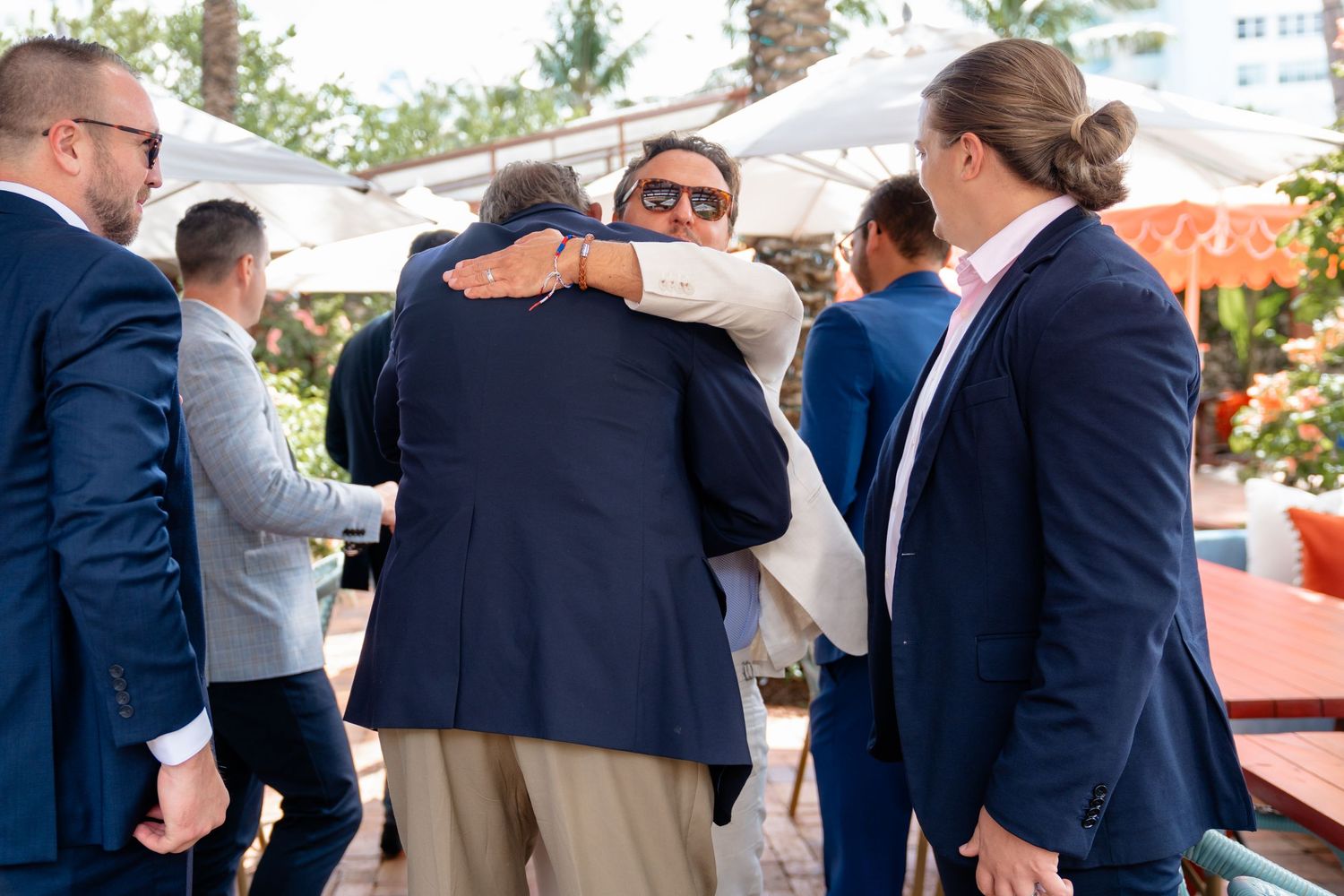 Men in suits embracing at an outdoor event, with others socializing in the background on a sunny day.