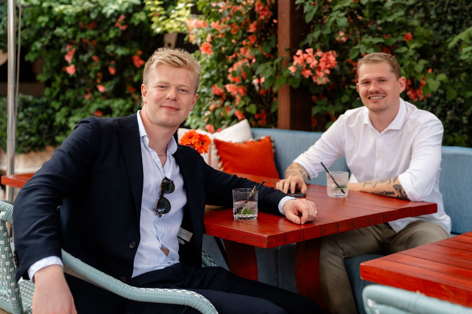 Two smiling men in smart casual attire sit at a red outdoor table with lush floral greenery backdrop.