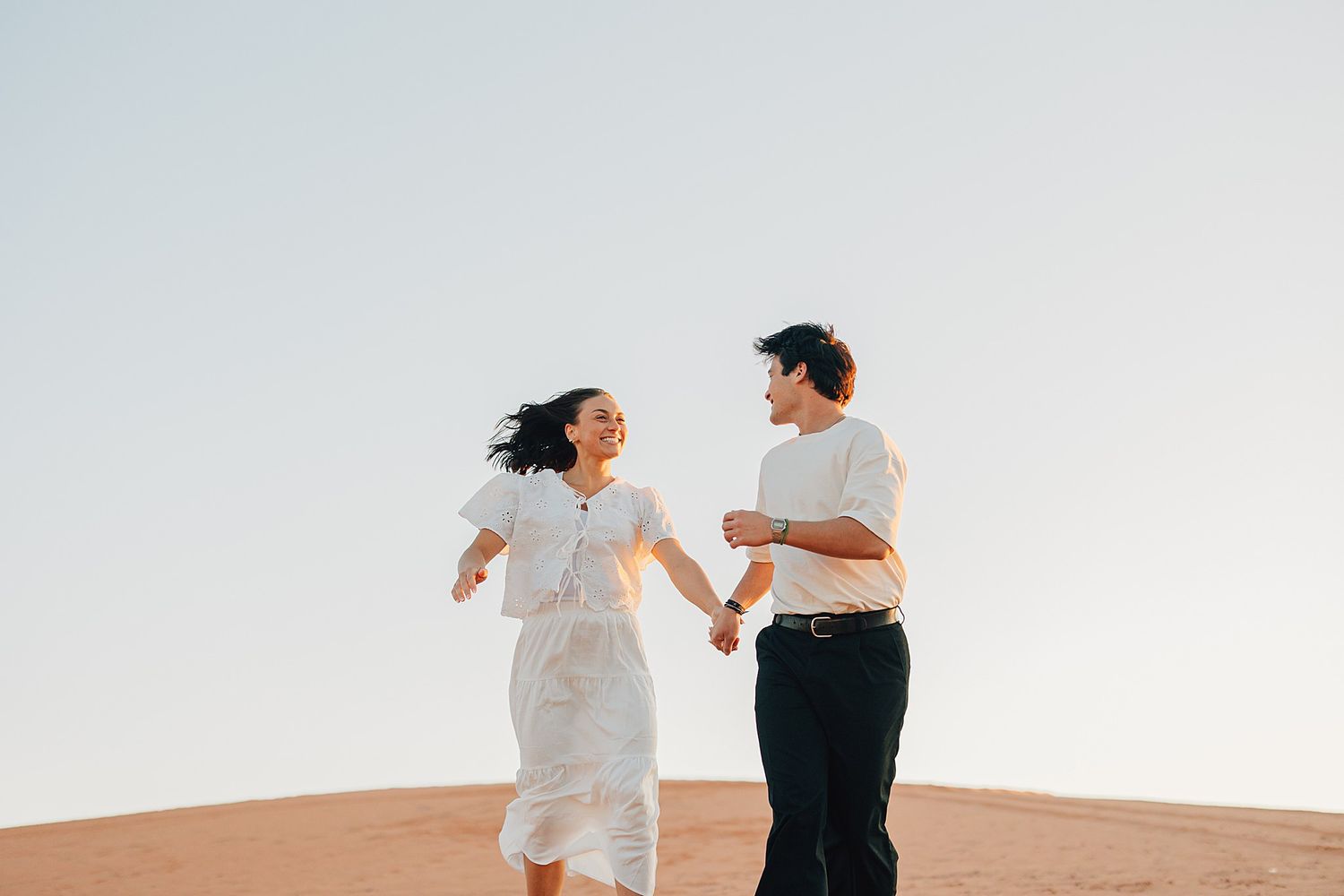 A joyful couple holds hands and runs together on a sandy desert dune under a clear sky.