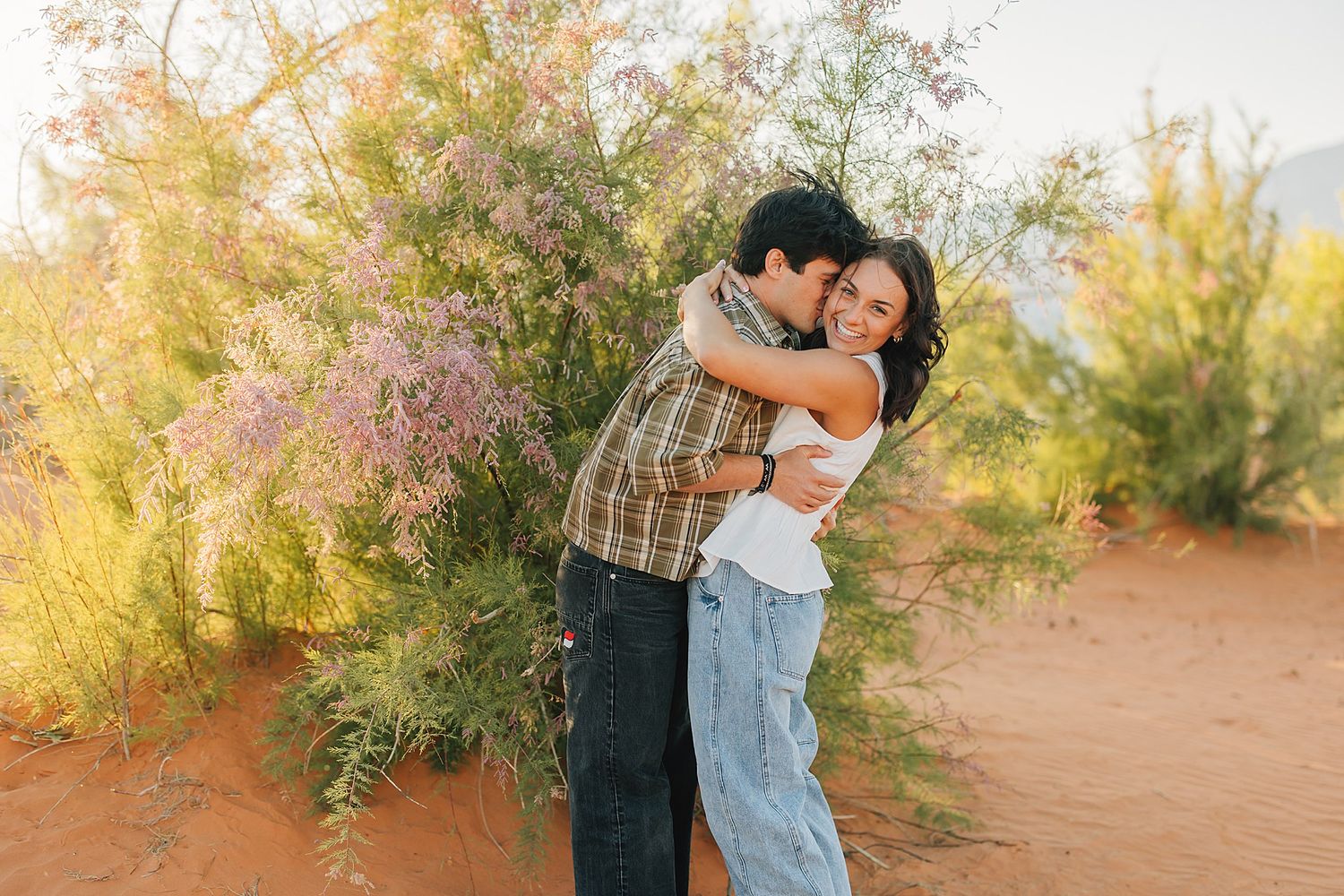 Couple embracing and smiling in a desert landscape with blooming pink shrubs and warm golden light.