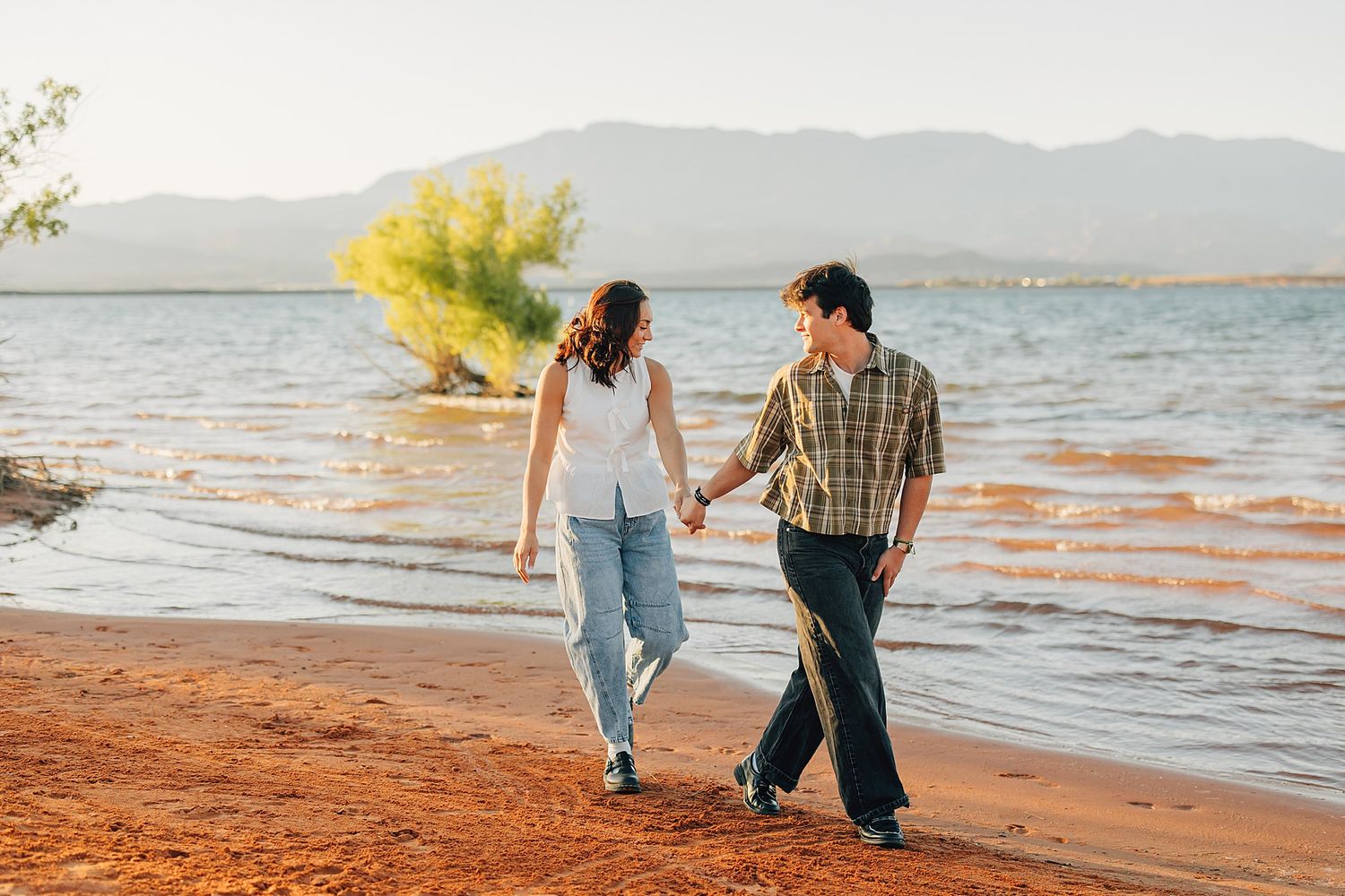 Couple holding hands walking along a scenic lakeside beach with mountains and green trees in the background.