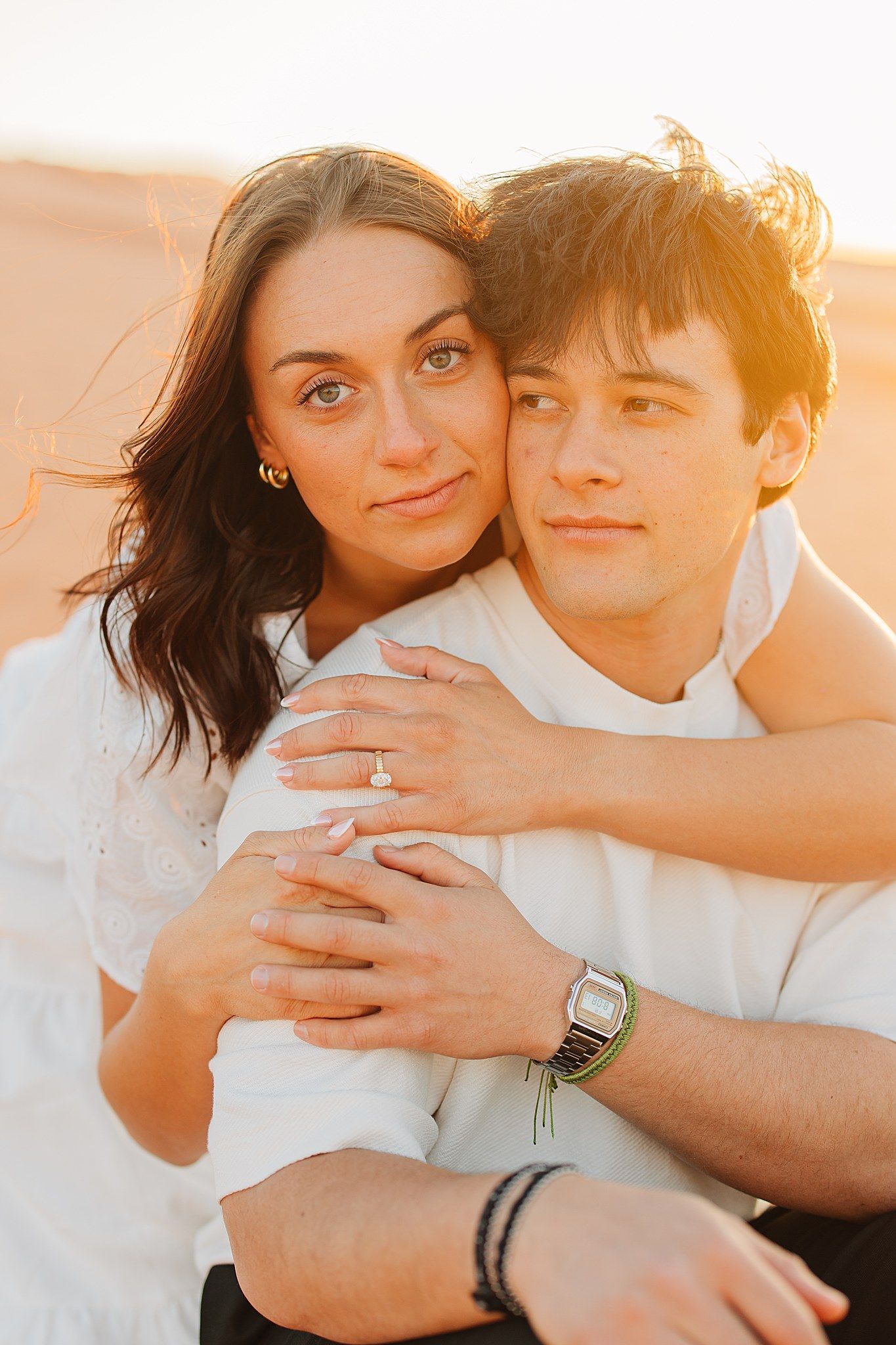 Couple embracing at sunset in a desert, woman with dark hair hugging man from behind, both wearing white shirts.