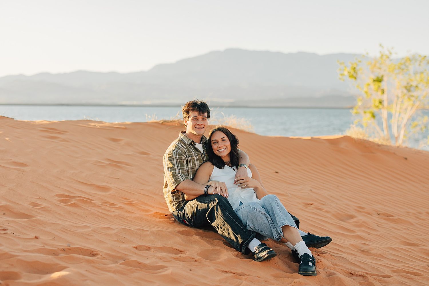 Couple sitting together on red sand dunes with scenic lake and mountains in background.
