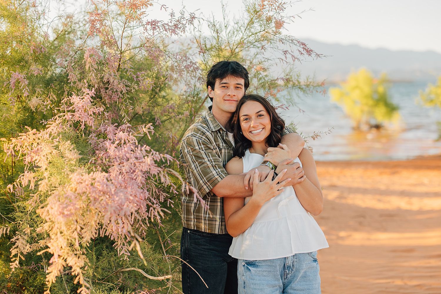 Couple embracing outdoors near blooming pink desert shrubs and a lake, smiling warmly in golden hour light.