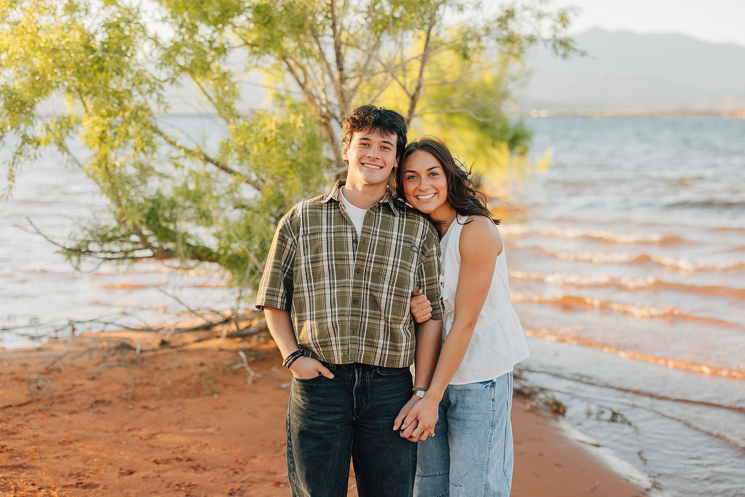 A smiling couple poses together near a lake shore with red sand, green trees, and mountains in the background.